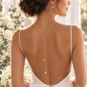 Op de afbeelding: Een goudkleurige rugketting met een fijne ketting die langs de ruggengraat valt, geaccentueerd met drie kleine witte parels op verschillende hoogtes. De ketting wordt gedragen bij een witte mouwloze jurk en bijpassende pareloorbellen.