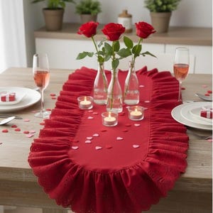May include: A red table runner with a ruffled edge, decorated with red roses in small glass vases, lit candles, and heart-shaped confetti. The table is set for a romantic dinner with champagne glasses and small gift boxes.