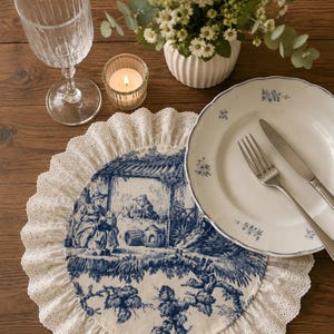 Op de afbeelding: Een ronde stoffen placemat met een blauw-wit toile de jouy-patroon met een centrale scène van figuren en gebladerte, afgewerkt met een brede witte kanten rand met oogjes. De placemat wordt getoond met een wit keramieken bord en bestek.