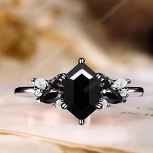 Könnte beinhalten: Ein silberner Ring mit einem großen, schwarzen, sechseckigen Mittelstein. Der Ring ist mit kleineren schwarzen und weißen Steinen verziert. Der Ring liegt auf einer hellen Oberfläche.