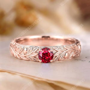 Op de afbeelding: Een roségouden ring met een ronde, gefacetteerde dieprode edelsteen in het midden. De band is voorzien van een doorlopend reliëfpatroon van fijne blaadjes en kleine besjes die de gehele omtrek van de metalen ring omsluiten.
