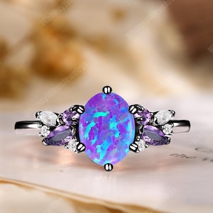 Könnte beinhalten: Ein silberner Ring mit einem großen, ovalen, blau-violetten Opal. Der Opal wird von kleineren lila und weißen Edelsteinen mit winzigen Akzentsteinen umgeben. Der Ring liegt auf einer hellen Oberfläche.