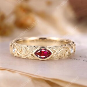 May include: A gold band ring featuring an engraved leaf pattern across the surface. The center of the ring is set with a marquise-cut red gemstone, flanked by two small round white diamonds on either side of the central stone setting.