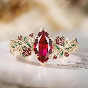 Puede incluir: Un anillo de oro con una gema roja oscura en forma de marquesa. La banda está adornada con diseños de rosas rojas y hojas verdes esmaltadas. El anillo está sobre una superficie blanca.