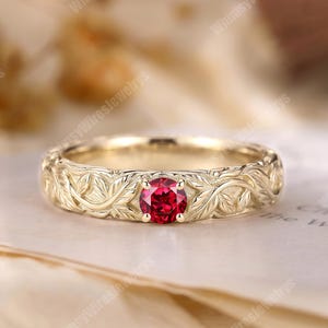 May include: A gold ring featuring a round, faceted red gemstone set in the center. The band is crafted with a detailed, raised relief pattern of intertwined leaves and vines that wraps around the entire circumference of the metal ring.