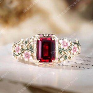 Op de afbeelding: Een gouden ring met een grote, rechthoekige, dieprode edelsteen in het midden. De band is versierd met verfijnde, gevormde metalen ranken, groene bladeren en kleine roze bloemen met witte harten, wat zorgt voor een gedetailleerd botanisch ontwerp.