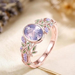 May include: A rose gold ring with an oval, light purple gemstone. The ring features a floral design with green leaves and purple flowers. The ring is set on a cream-coloured surface.