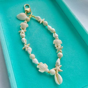 May include: A beach-themed keyring with a gold-coloured clasp and a strand of white seashells, starfish, turtle charms, and gold beads. The keyring is designed with a variety of shell shapes and sizes, creating a coastal aesthetic.
