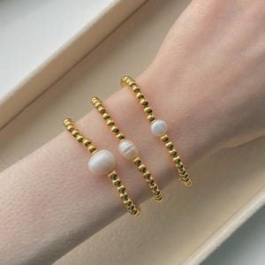 May include: Three gold beaded bracelets with white pearls. The bracelets are made of small, round gold beads and feature a single, large pearl or two smaller pearls. The bracelets are worn on a person's wrist.