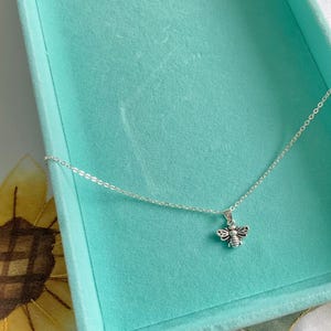 May include: A silver necklace featuring a bee pendant. The bee charm has detailed wings and body segments. The necklace is presented on a turquoise surface, with a sunflower visible in the background.