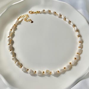 May include: A necklace made of white seashells and gold-coloured beads. The necklace has a gold-coloured clasp and is displayed on a white surface. The seashells are strung together with small gold-coloured beads.