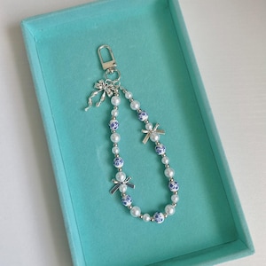 May include: A beaded keychain featuring white faux pearls and blue and white ceramic beads. The keychain has silver-tone accents, including bow-shaped charms and a clasp. The keychain is presented in a turquoise box.