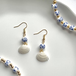 May include: Gold-coloured earrings featuring a blue floral bead and a white seashell charm. Part of a jewellery set with a matching bracelet. The jewellery is displayed on a white surface.