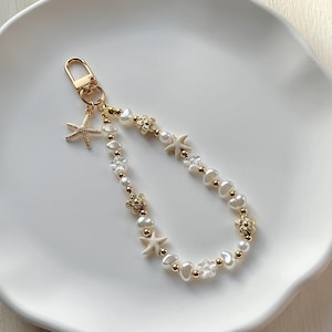 May include: A gold-tone keychain with a lobster clasp, featuring a strand of white pearls, gold beads, and decorative charms. The strand includes starfish and flower-shaped elements. The keychain has a beach or coastal theme.