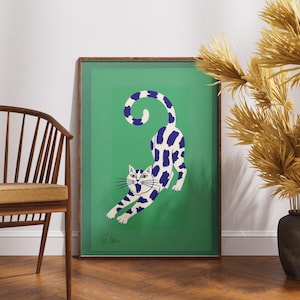 May include: A framed art print featuring a stylized cat stretching. The cat is white with blue spots and a blue and white striped tail, set against a green background. The frame is a dark wood color. The artwork is displayed near a wooden chair and decorative dried plants.