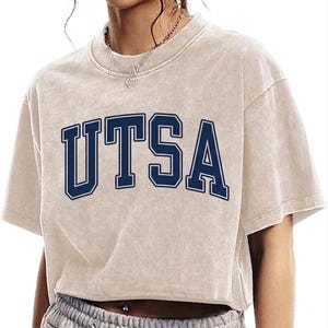 May include: A beige crop top with the word "UTSA" in navy blue block letters. The shirt has short sleeves and a slightly faded appearance. The overall style is casual and relaxed.