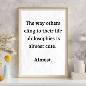 May include: A framed print with the quote, "The way others cling to their life philosophies is almost cute. Almost." The text is in black on a white background, framed in gold. The print is displayed on a white countertop.