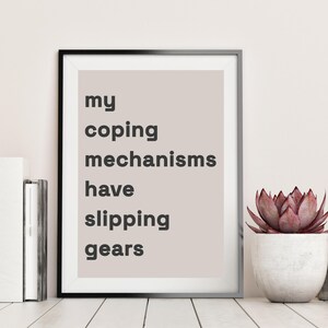 Könnte beinhalten: Gerahmter Druck mit dem Text "my coping mechanisms have slipping gears" in schwarzer Schrift auf hellbeigem Hintergrund. Der Druck in einem schwarzen Rahmen steht auf einer weißen Oberfläche neben einer Sukkulente in einem weißen Topf.