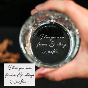 Puede incluir: Un vaso transparente con las palabras "I love you more forever & always Alex" grabadas en la base. El vaso lo sostiene una persona. Una pequeña tarjeta blanca con el mismo texto está en la esquina inferior izquierda.