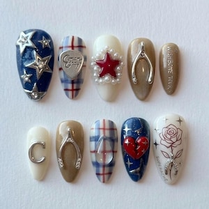 May include: A set of ten artificial nails with various designs. The designs include stars, hearts, plaid, a wishbone, and a rose. Colors include blue, red, white, and brown. Some nails feature embellishments like pearls and silver charms. The text "Conan Gray" and "Wishbone Tours" are visible.