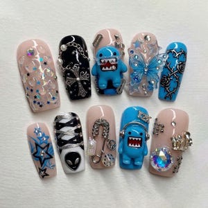 May include: A set of ten artificial nails with various designs. The nails feature a range of colors, including pink, black, and blue, and are adorned with embellishments such as stars, chains, and a cartoon character. Some nails have a glossy finish.