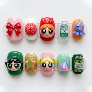 May include: A set of ten artificial nails with various 3D designs. The designs include a red bow, a brain, a Powerpuff Girl, a vial with "CHEMICAL X", an octopus, a star, a flask, and a jar of "MAYOR'S PICKLES".