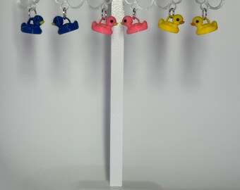May include: Earrings featuring small, colorful rubber duck charms. The earrings are in blue, pink, and yellow, with silver-colored hooks. The charms are hanging from clear, circular earring displays.