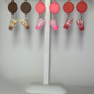 May include: A collection of six pairs of earrings. The earrings feature bottle cap tops in brown, pink, and red, with miniature milkshake charms. The milkshake charms are in shades of brown, pink, and red, with white accents.