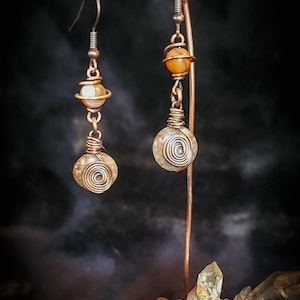 Puede incluir: Pendientes artesanales de cobre con dise&ntilde;o en espiral. Cada pendiente presenta una peque&ntilde;a piedra redonda y clara, y una piedra m&aacute;s grande envuelta en espiral, suspendidas de un gancho de cobre. Se exhiben en un soporte de cobre.