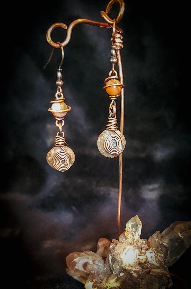 Puede incluir: Pendientes artesanales de cobre con dise&ntilde;o en espiral. Cada pendiente presenta una peque&ntilde;a piedra redonda y clara, y una piedra m&aacute;s grande envuelta en espiral, suspendidas de un gancho de cobre. Se exhiben en un soporte de cobre.