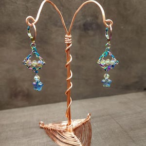 May include: A pair of handmade earrings displayed on a copper wire stand. The earrings feature a diamond-shaped frame with iridescent accents, clear beads, and a blue star-shaped charm. The stand has a twisted stem and a triangular base.