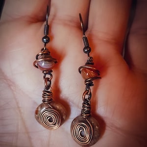 Puede incluir: Pendientes hechos a mano con cuentas de piedra natural y detalles de alambre de cobre. Los pendientes tienen un dise&ntilde;o en espiral en la piedra inferior, con un envoltorio de alambre de cobre alrededor de la piedra del medio.