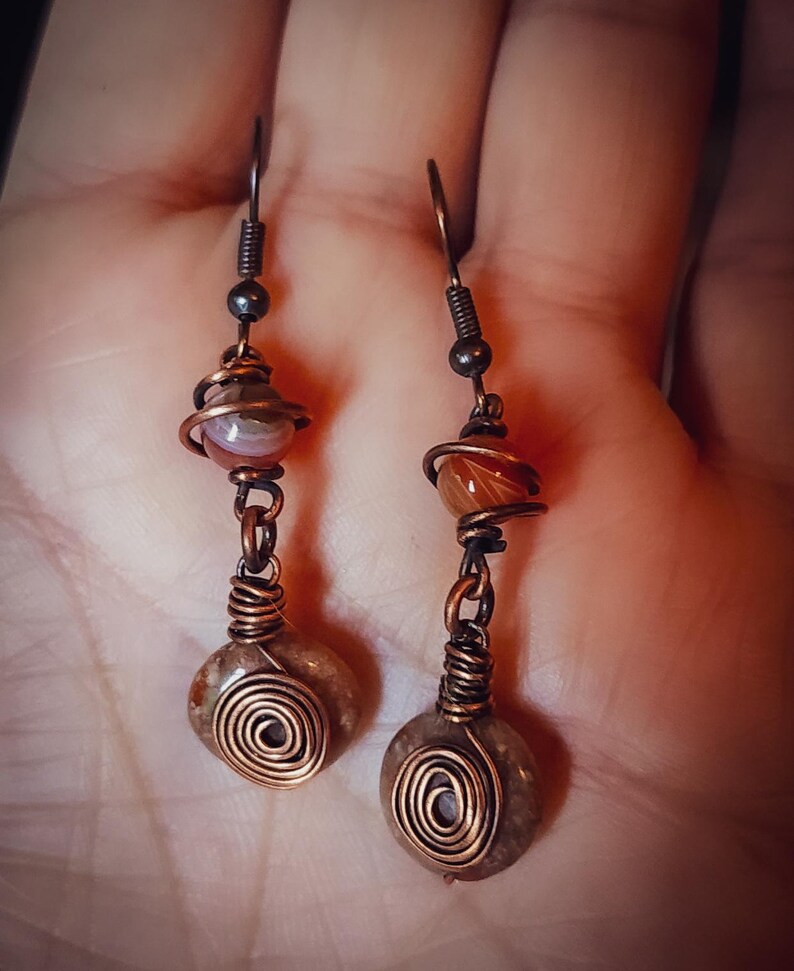 Puede incluir: Pendientes hechos a mano con cuentas de piedra natural y detalles de alambre de cobre. Los pendientes tienen un dise&ntilde;o en espiral en la piedra inferior, con un envoltorio de alambre de cobre alrededor de la piedra del medio.