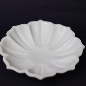 May include: A white, petal-shaped ceramic dish with a matte finish. The dish has a shallow bowl and a scalloped edge, resembling a flower. The background is a solid black.