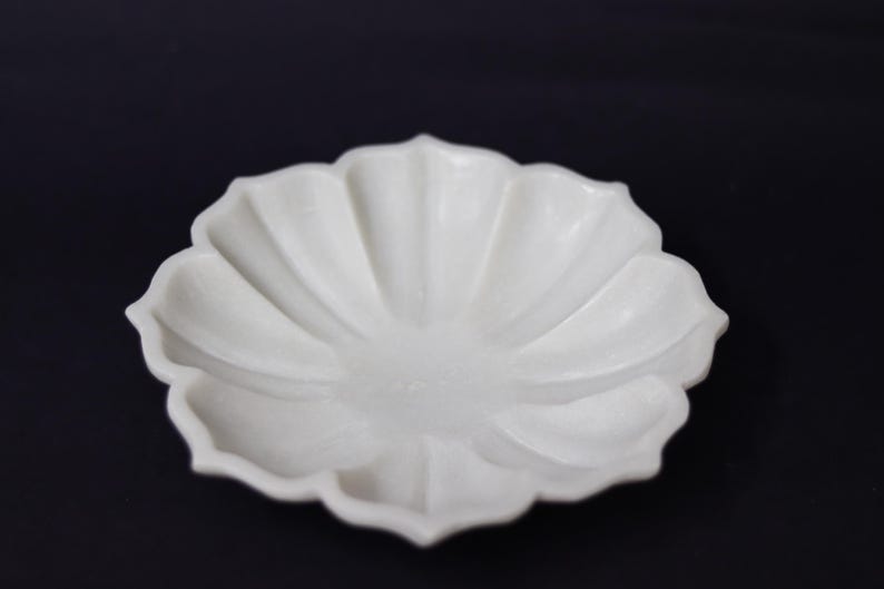 May include: A white, petal-shaped ceramic dish with a matte finish. The dish has a shallow bowl and a scalloped edge, resembling a flower. The background is a solid black.