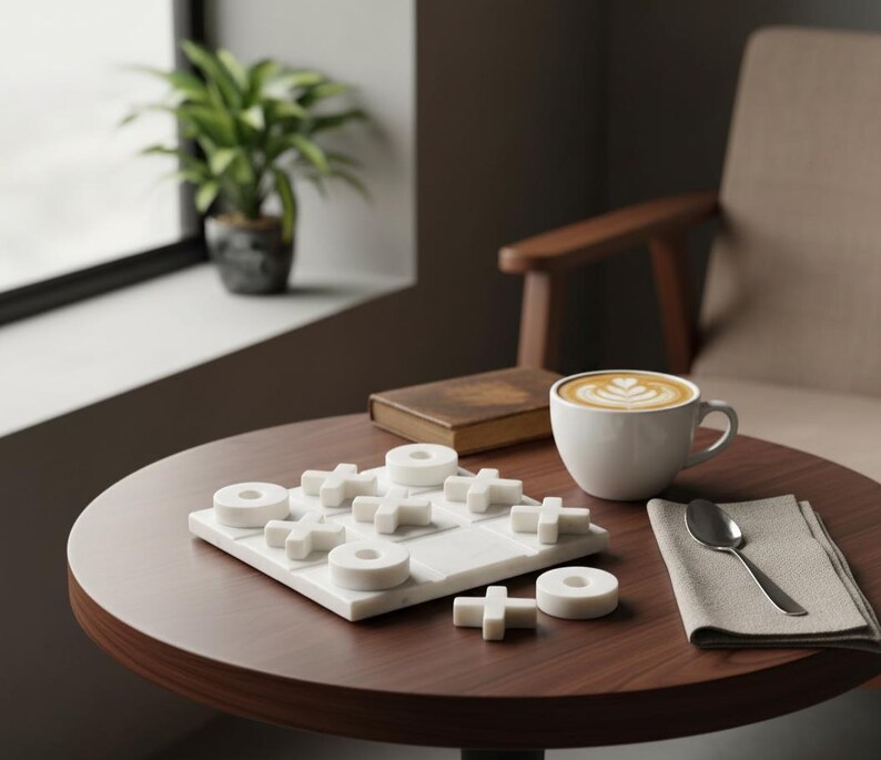 May include: A round, dark brown table holds a white marble tic-tac-toe game, a white coffee cup with latte art, a spoon, a linen napkin, and a small, brown book. A potted plant sits near a window. A beige armchair is in the background.