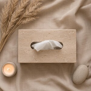 May include: A rectangular beige stone tissue box cover with a textured, porous surface. A single white tissue is pulled through the central oval opening. The box features natural mineral variations in a light tan color, providing a solid, structured appearance for holding standard facial tissues.
