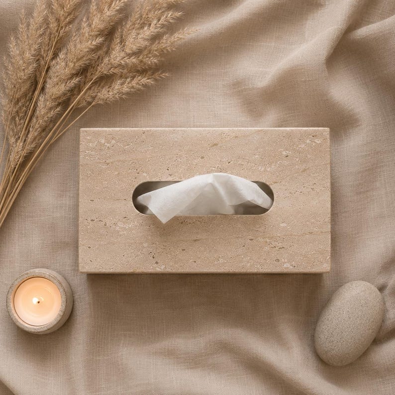 May include: A rectangular beige stone tissue box cover with a textured, porous surface. A single white tissue is pulled through the central oval opening. The box features natural mineral variations in a light tan color, providing a solid, structured appearance for holding standard facial tissues.