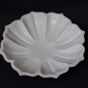 May include: A white, flower-shaped decorative dish with a scalloped edge. The dish has a smooth, matte finish and is set against a black background. The dish's design features a central depression and radiating petal-like ridges.
