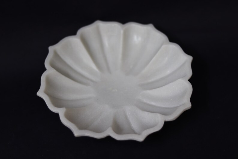 May include: A white, flower-shaped decorative dish with a scalloped edge. The dish has a smooth, matte finish and is set against a black background. The dish's design features a central depression and radiating petal-like ridges.