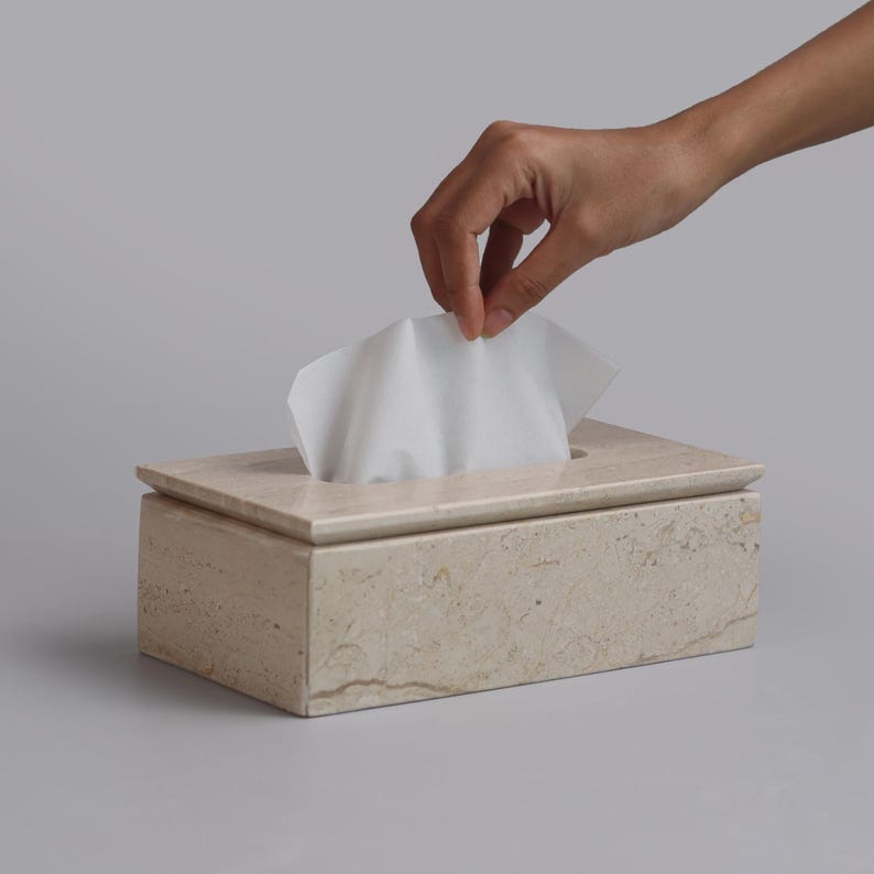 May include: A rectangular tissue box cover crafted from beige travertine stone with a natural porous texture and a smooth finish. The box features a central rectangular opening on the top surface, through which a single white paper tissue is pulled.