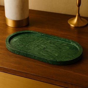 Puede incluir: Una bandeja ovalada de mármol verde con borde elevado. La bandeja tiene un patrón veteado natural y se exhibe sobre una superficie de madera. La bandeja mide aproximadamente 30 cm de largo.