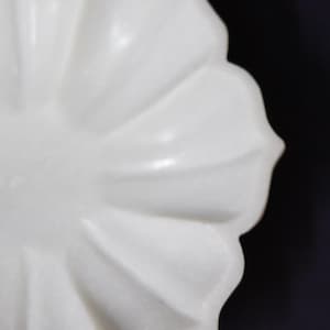 May include: A white, petal-shaped decorative bowl. The bowl has a smooth, glossy finish and a scalloped edge. The design resembles a flower, with each petal clearly defined. The background is a dark, solid color.