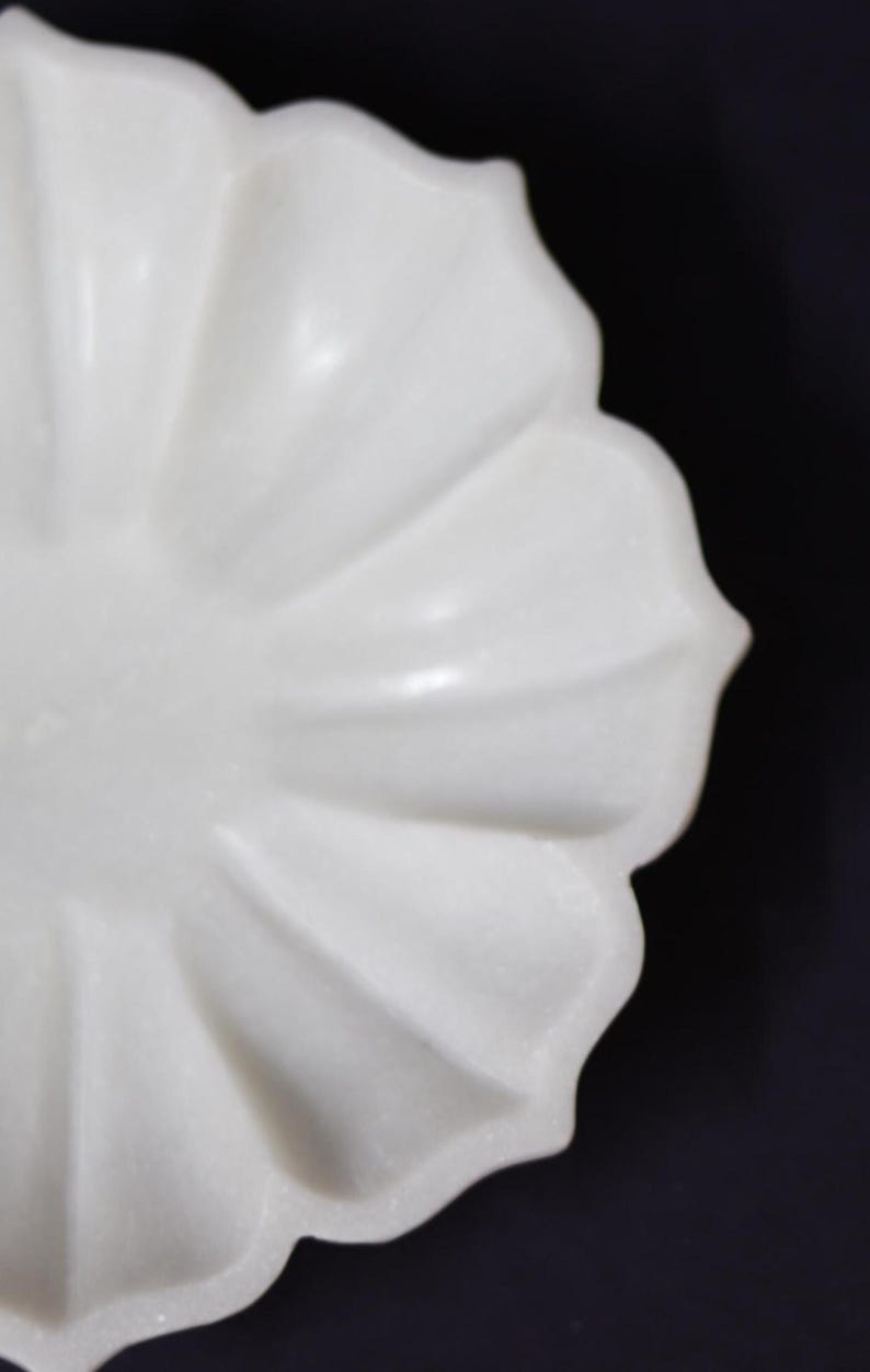 May include: A white, petal-shaped decorative bowl. The bowl has a smooth, glossy finish and a scalloped edge. The design resembles a flower, with each petal clearly defined. The background is a dark, solid color.