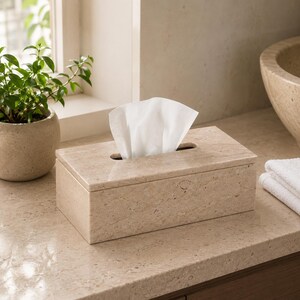 May include: A rectangular tissue box cover crafted from polished beige stone with a natural speckled pattern. The box features a centered oval opening at the top, through which a single white facial tissue is pulled upward for easy access.
