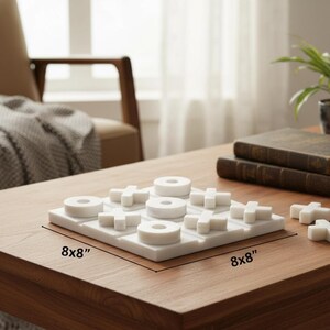 May include: A white marble tic-tac-toe game set. The game board is 8x8 inches, with circular and cross-shaped game pieces. The set is displayed on a wooden table, with books and a potted plant in the background.