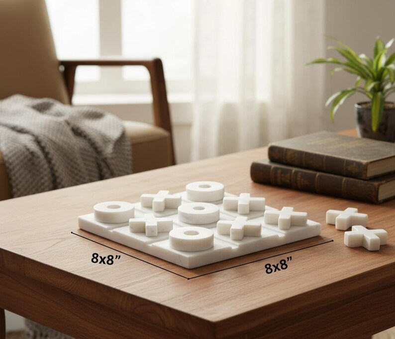 May include: A white marble tic-tac-toe game set. The game board is 8x8 inches, with circular and cross-shaped game pieces. The set is displayed on a wooden table, with books and a potted plant in the background.