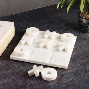 May include: A white marble tic-tac-toe game set. The game board features a grid with carved lines, and the game pieces are white X's and O's. The set is displayed on a dark surface, with a book and a potted plant in the background.