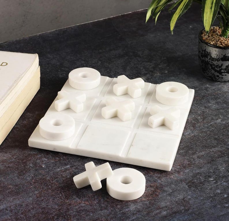 May include: A white marble tic-tac-toe game set. The game board features a grid with carved lines, and the game pieces are white X's and O's. The set is displayed on a dark surface, with a book and a potted plant in the background.