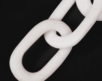 May include: A close-up of a white chain against a black background. The chain links are oval-shaped and interlocked, creating a pattern. The chain has a smooth, matte finish. The image is well-lit, highlighting the chain's texture and form.
